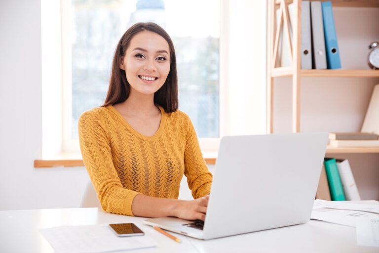 Cheerful businesswoman dressed in sweater sitting in office while using laptop and looking at camera