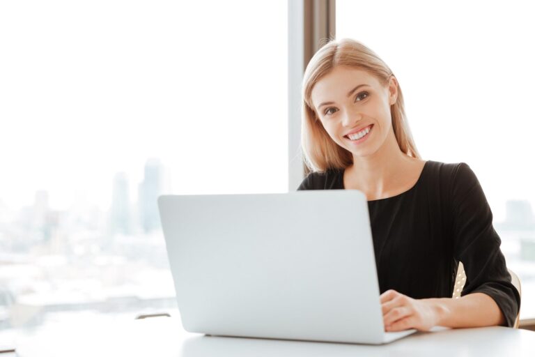 Picture of cheerful young lady worker sitting in office while using laptop computer and typing by keyboard. Look at camera.